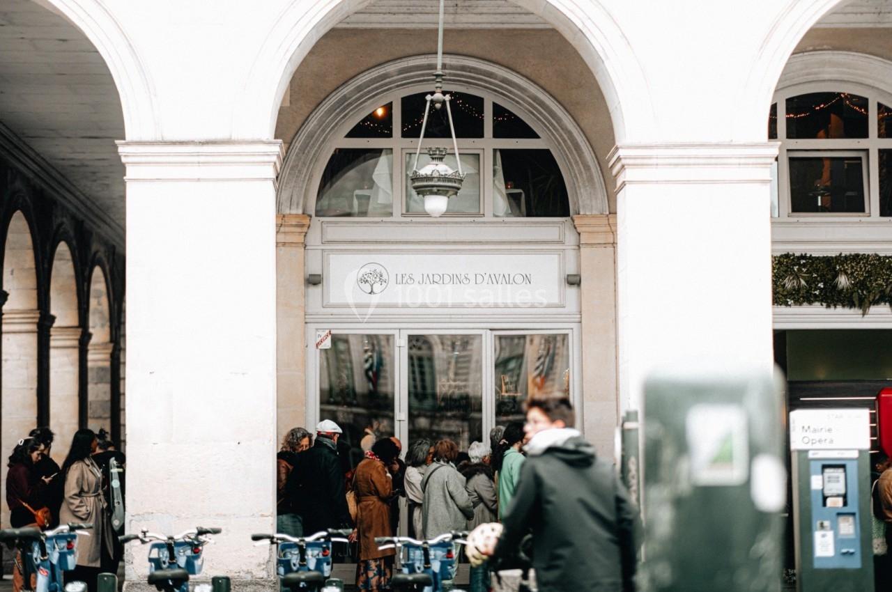Entrée d'un bâtiment avec une enseigne ’Les Jardins d'Avallon’, des personnes rassemblées devant et des vélos stationnés.