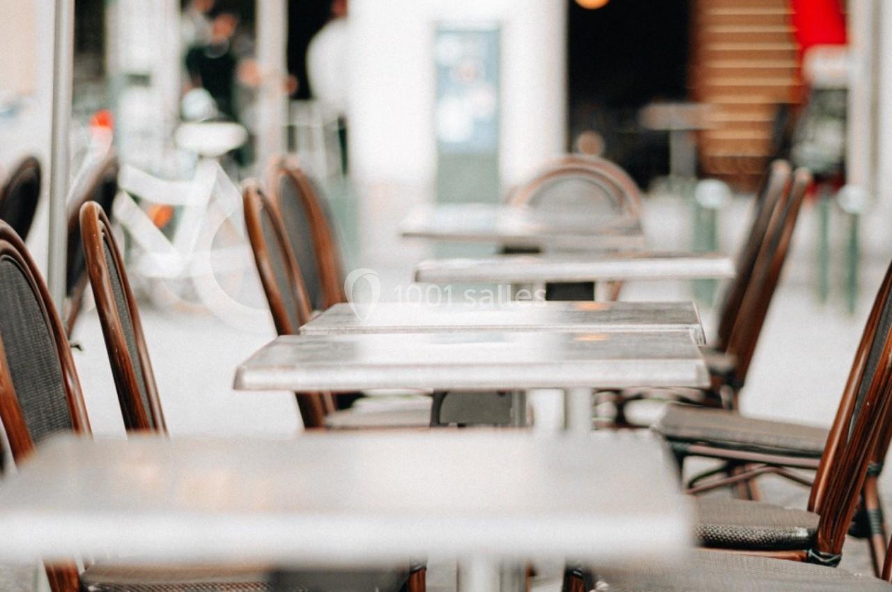Terrasse de café avec des tables et chaises en osier vides, partiellement ombragée par des parasols.