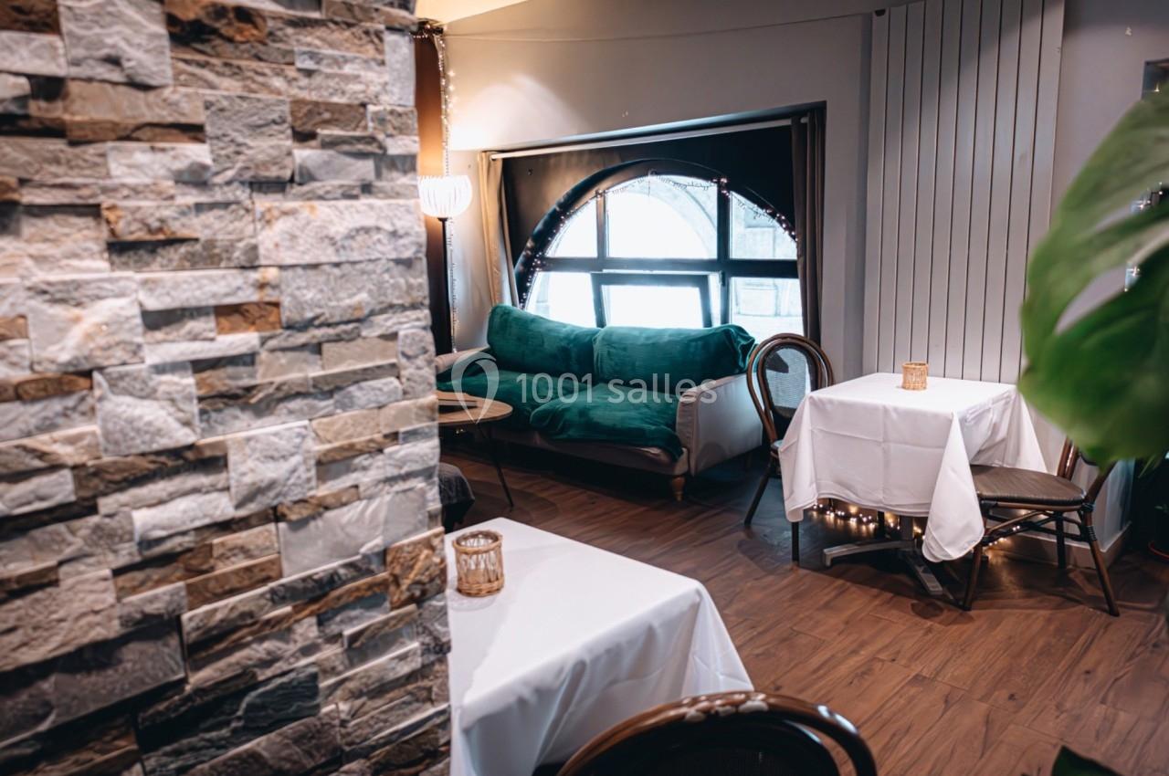 Salle de restaurant cosy avec tables nappées, fauteuils verts, mur en pierre et grande fenêtre arrondie.