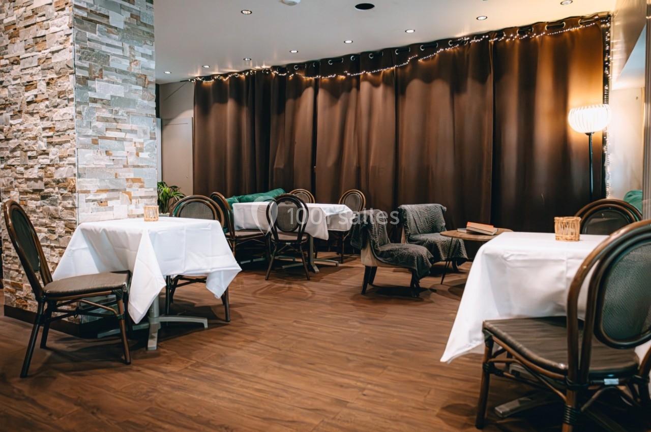 Salle de restaurant avec tables dressées, chaises en rotin, sol en bois et rideaux sombres en arrière-plan.