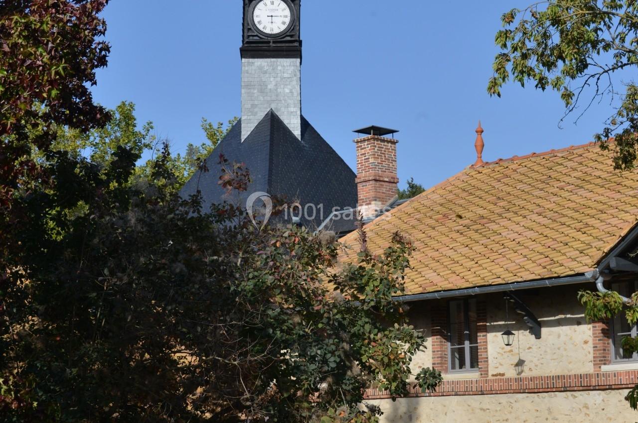 Clocher d'église avec horloge visible, entouré de bâtiments en briques et de végétation sous un ciel bleu.