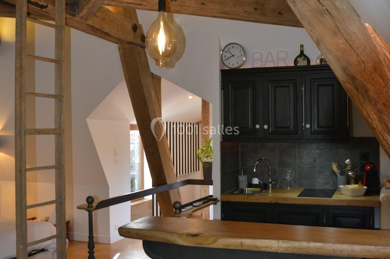 Cuisine ouverte dans un intérieur avec poutres apparentes, tabourets hauts, et éclairage suspendu.