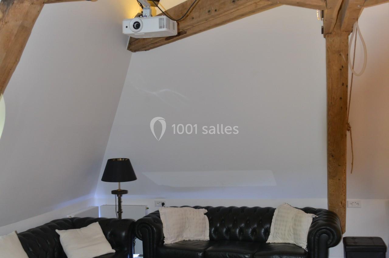 Salon mansardé avec poutres apparentes, canapés en cuir noir, table basse originale et projecteur au plafond.