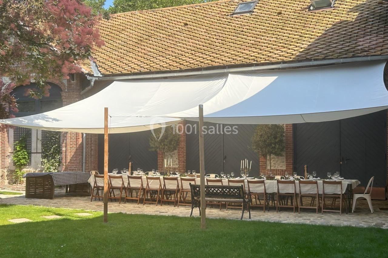 Grande table dressée sous des voiles d'ombrage blanches dans une cour pavée, devant un bâtiment en briques.