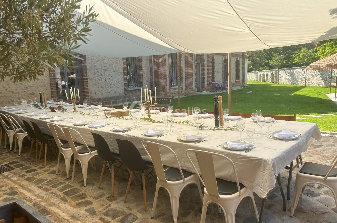 Grande table extérieure dressée avec nappes blanches, chaises variées et décorations, sous une toile d'ombrage.