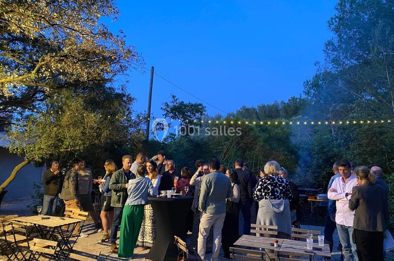 Groupe de personnes discutant dans un jardin en soirée, entouré de tables et éclairé par des guirlandes lumineuses.