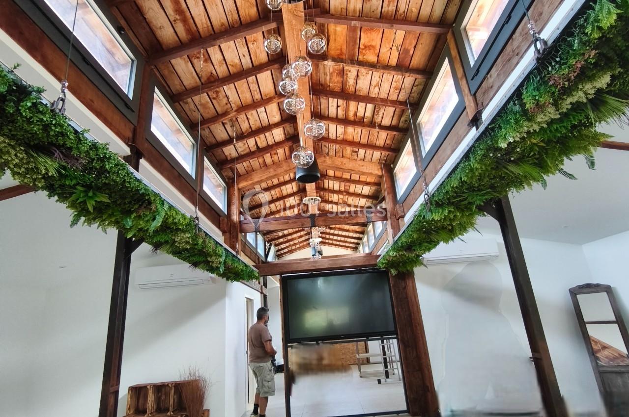 Plafond en bois avec poutres apparentes, rangées de plantes suspendues et un écran au centre d'une pièce lumineuse.