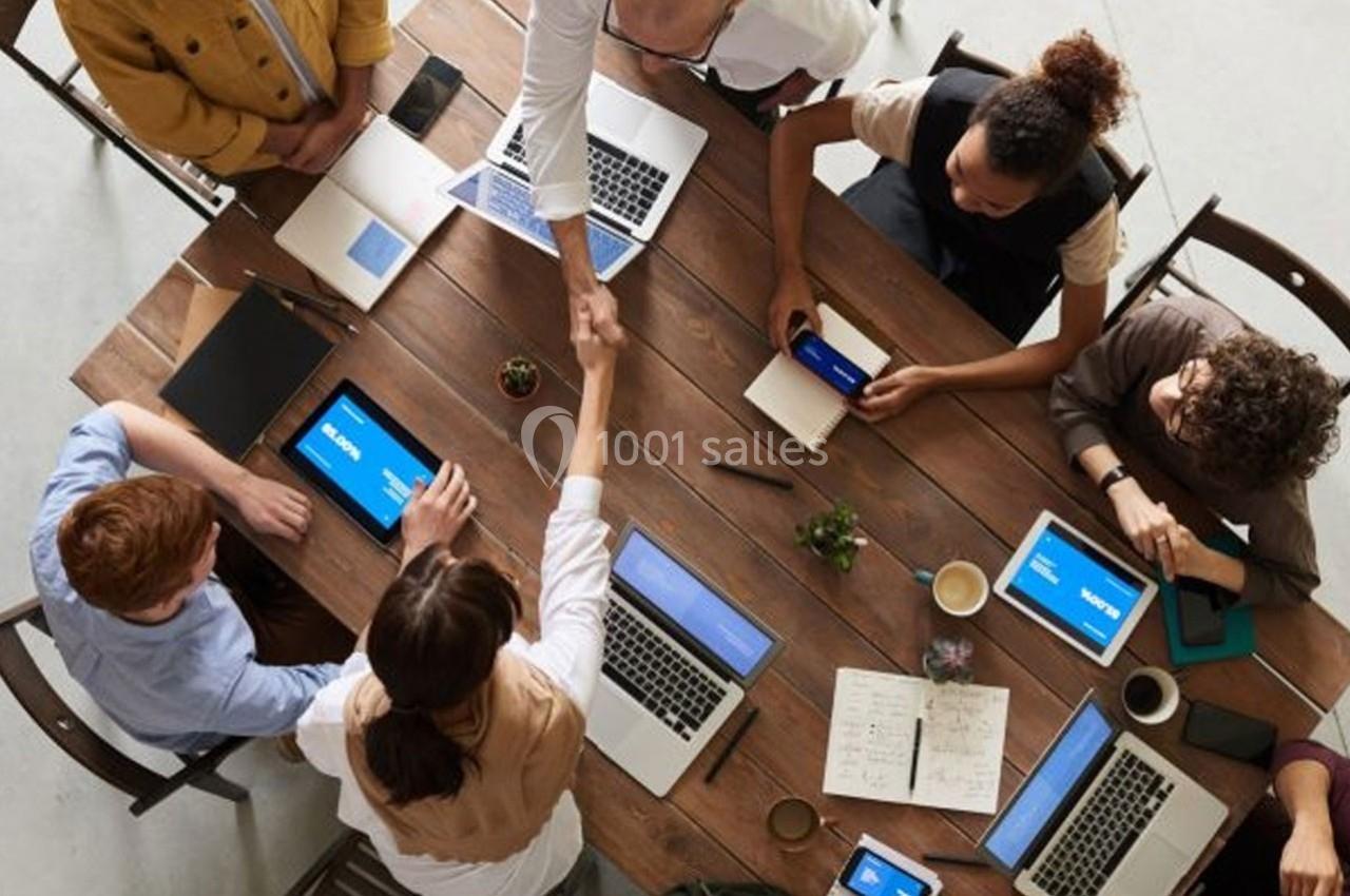 Personnes collaborant autour d'une table avec ordinateurs, tablettes, carnets et une poignée de main au centre.