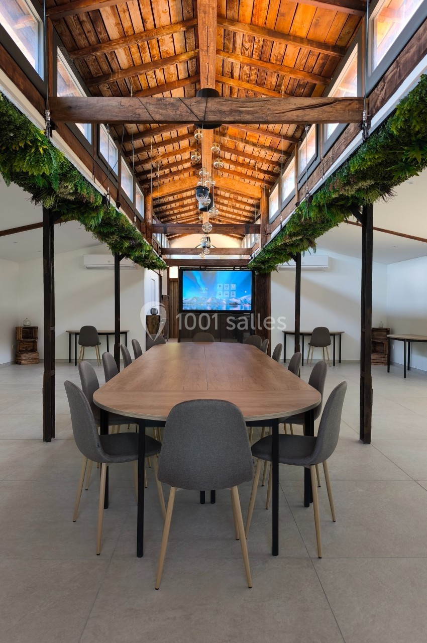 Salle de réunion lumineuse avec une grande table en bois entourée de chaises grises, sous un toit en bois avec plantes…