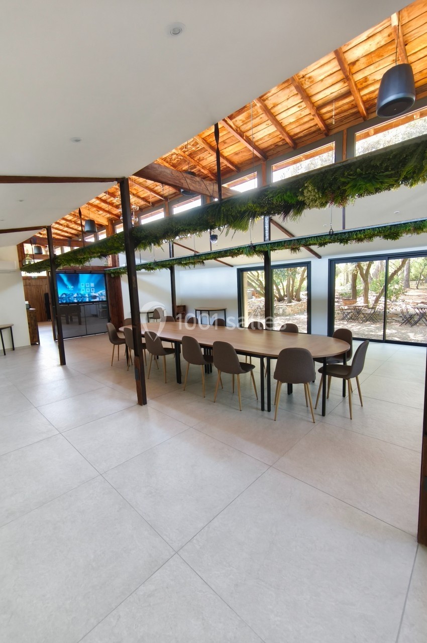 Grande salle lumineuse avec une longue table en bois entourée de chaises, toit en bois et grandes baies vitrées donnant sur…