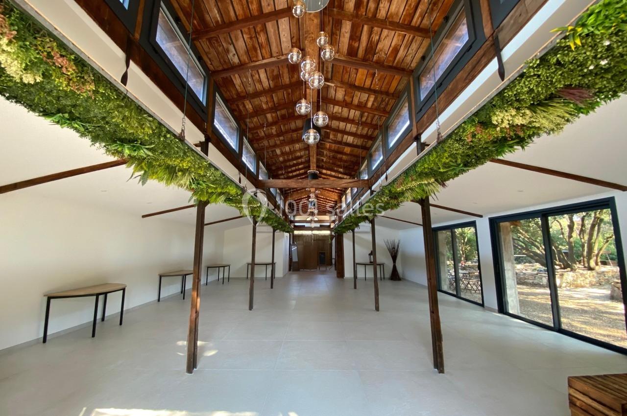 Salle lumineuse avec charpente en bois, suspensions modernes, murs blancs et grandes baies vitrées donnant sur un jardin.