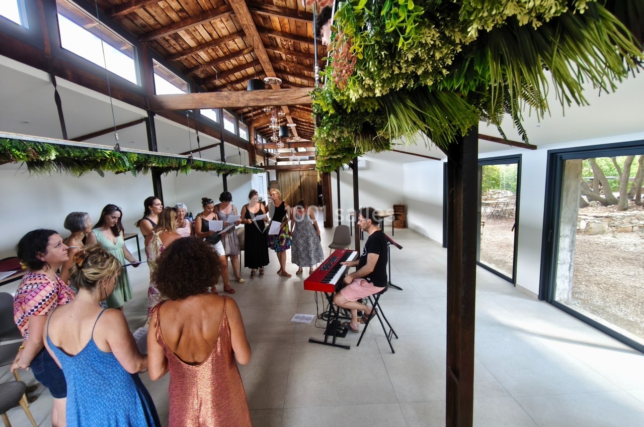 Un groupe de personnes chante autour d'une pianiste dans une salle lumineuse avec un plafond en bois et des plantes…