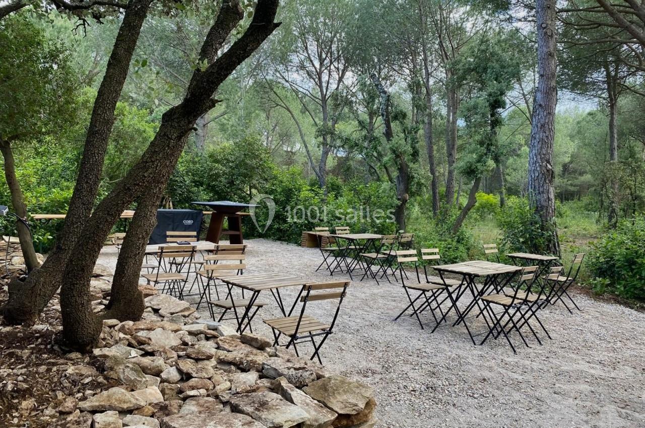 Tables et chaises en métal et bois disposées sur un sol gravillonné, entourées d'arbres dans un cadre naturel.