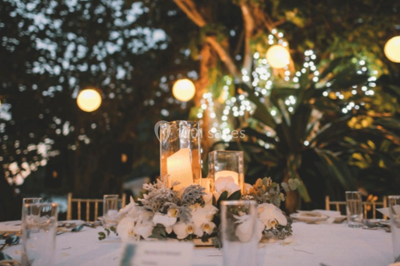 Table décorée avec des bougies, fleurs blanches et verres, entourée de guirlandes lumineuses dans un jardin en soirée.