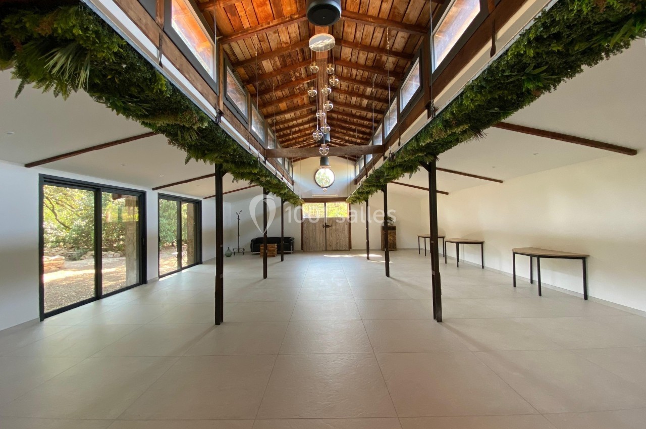 Salle spacieuse avec toit en bois, grandes baies vitrées, tables le long des murs et décoration végétale suspendue.