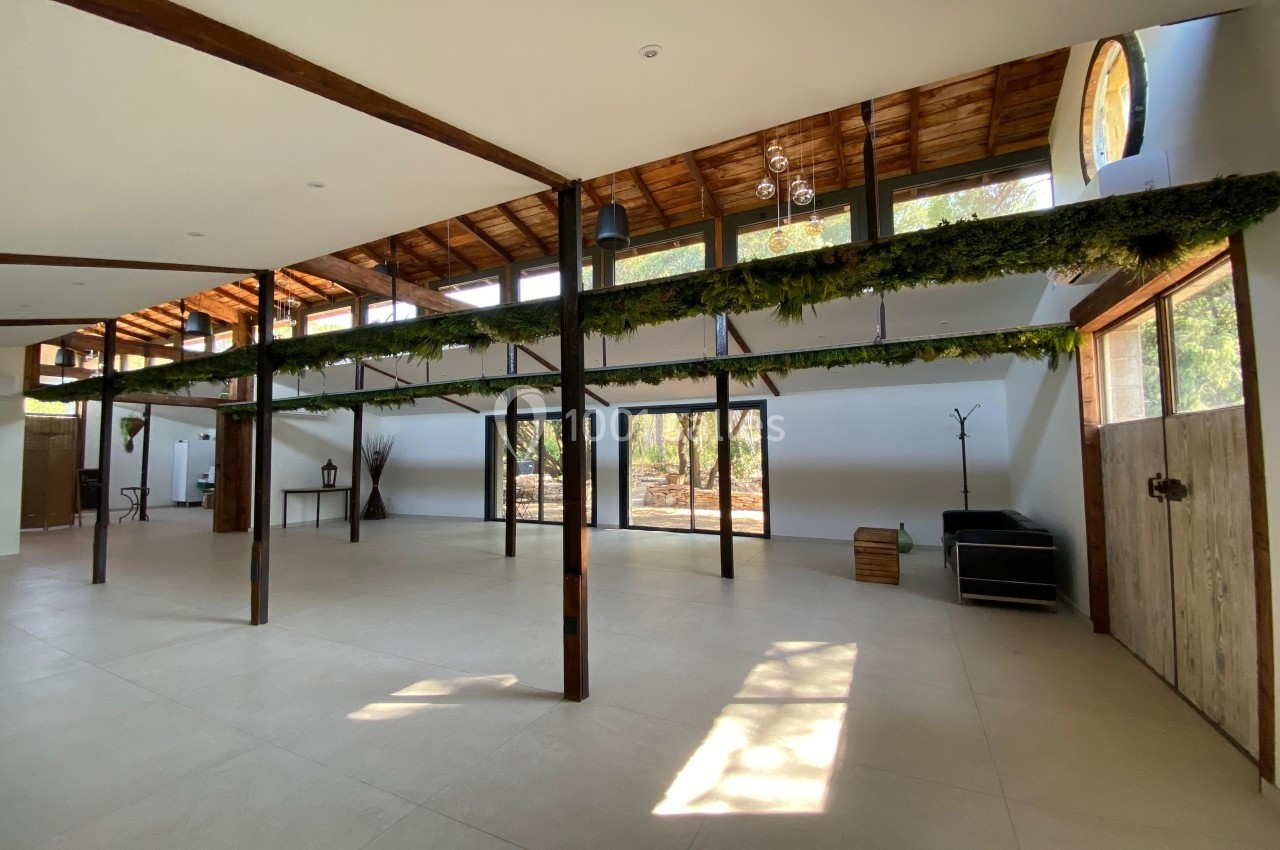 Intérieur spacieux avec poutres en bois, grandes baies vitrées, plafond en pente et décoration végétale suspendue.