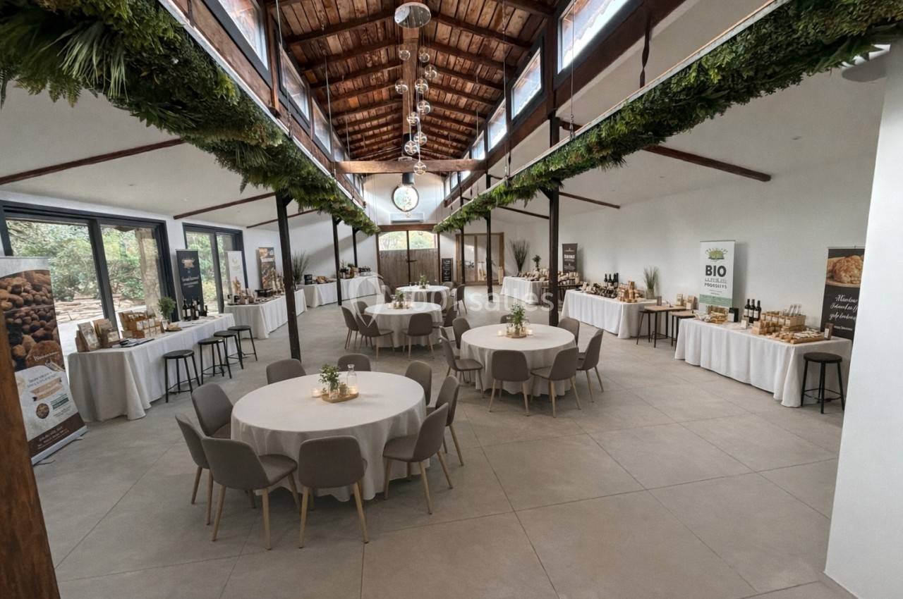 Salle lumineuse avec tables rondes dressées, décorations végétales suspendues et stands de produits exposés.