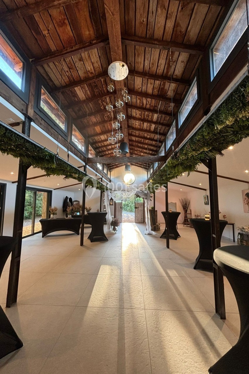 Salle lumineuse avec charpente en bois, décorée de guirlandes végétales, tables hautes noires et grandes baies vitrées.