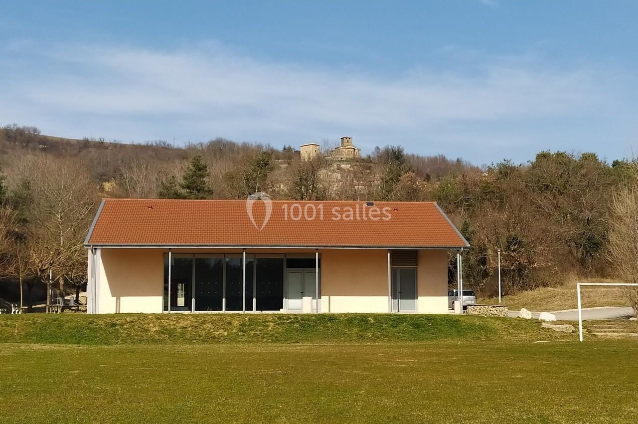 Bâtiment avec toit rouge et façade beige, situé devant une colline boisée et un terrain de sport.