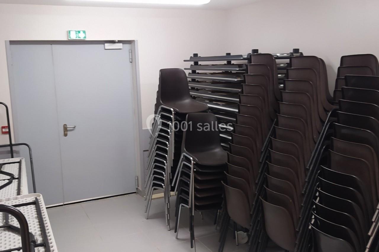 Salle de stockage avec chaises empilées, tables pliées et une porte grise équipée d'une sortie de secours.