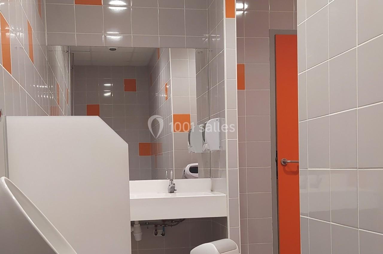 Lavabo blanc avec miroir dans des toilettes carrelées de gris et orange, porte orange visible à droite.