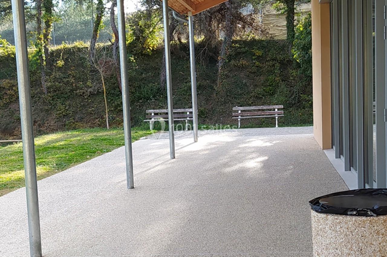 Terrasse couverte avec sol en béton, vue sur un espace vert arboré et deux bancs en arrière-plan.