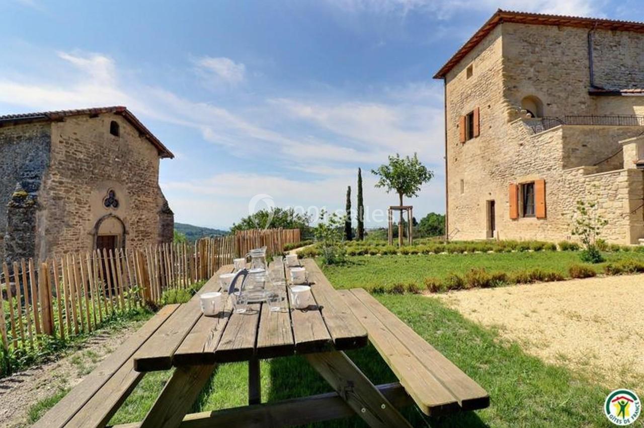 Table de pique-nique en bois dans un jardin, entourée de bâtiments en pierre et d'une clôture en bois.