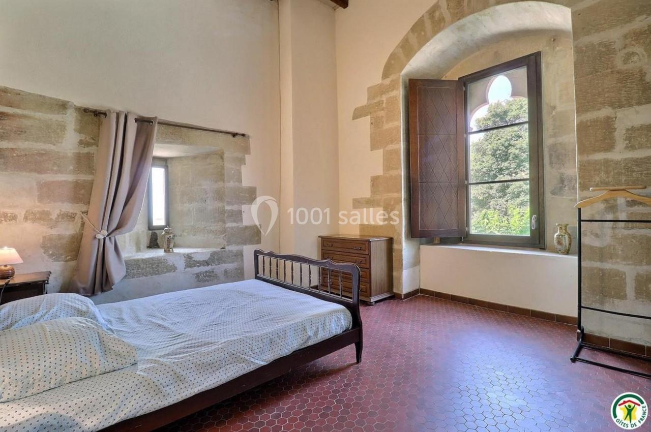 Chambre lumineuse avec lit en métal, murs en pierre, sol en carrelage rouge et grande fenêtre donnant sur la verdure.