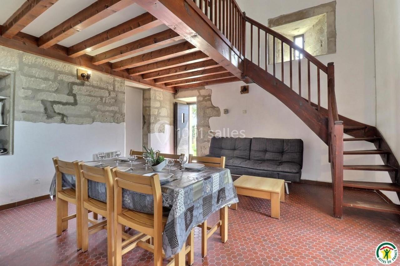 Salle à manger avec table dressée, canapé, escalier en bois et murs en pierre apparente dans un intérieur lumineux.