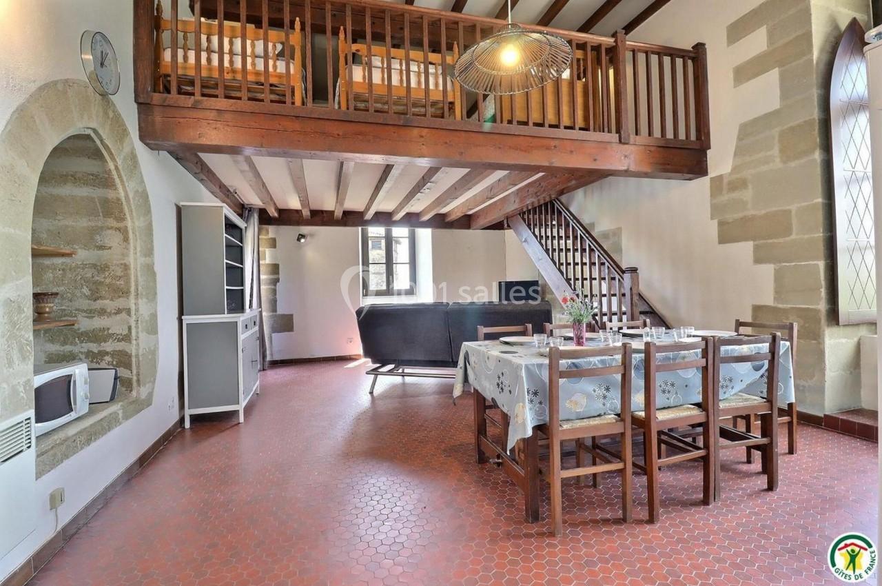 Salle à manger avec table en bois, chaises, mezzanine en bois, murs en pierre et grande fenêtre lumineuse.