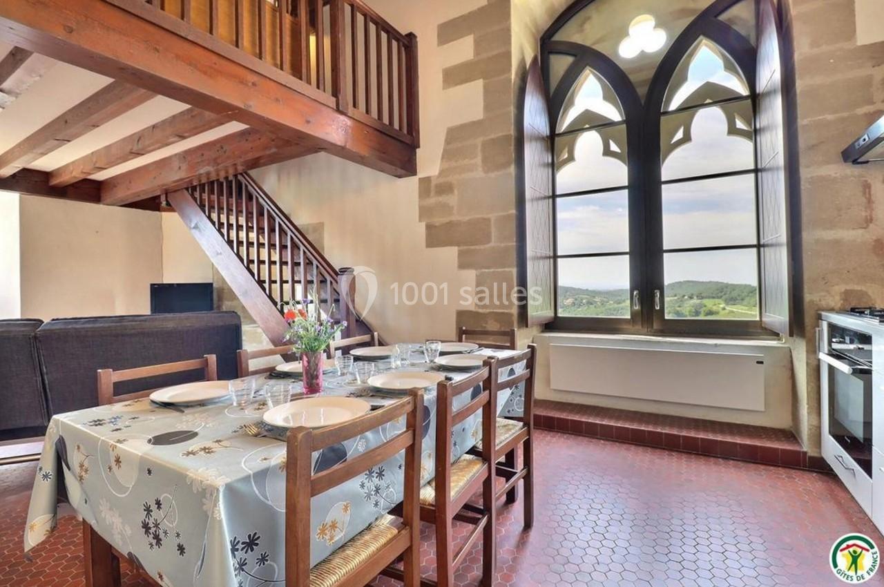 Salle à manger lumineuse avec table dressée, escalier en bois et grande fenêtre offrant une vue sur la campagne.