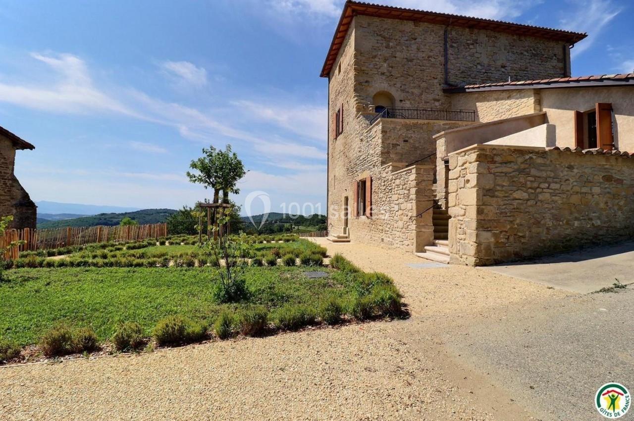Maison en pierre avec terrasse, entourée de verdure et d'un jardin, sous un ciel dégagé en milieu rural.