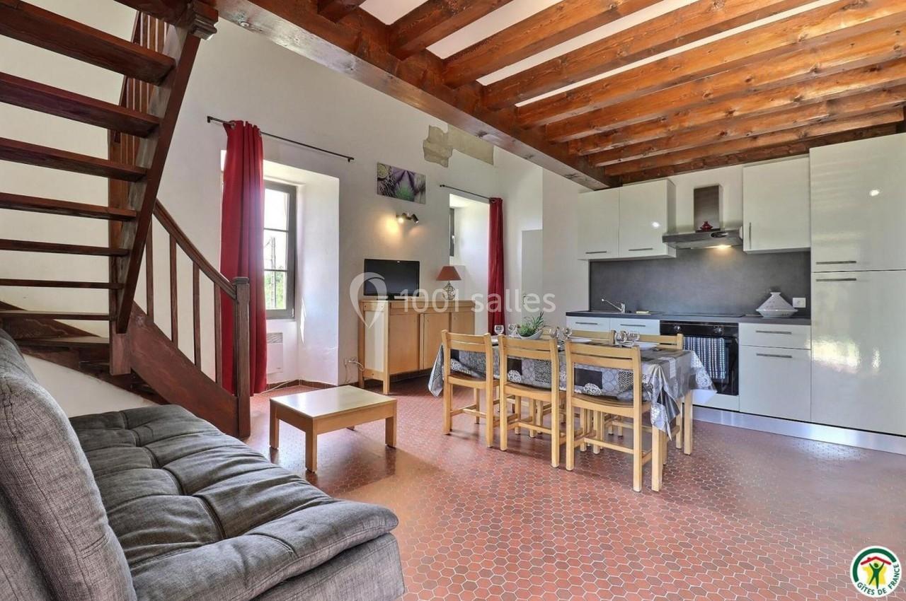 Salon avec canapé, table à manger, cuisine équipée et escalier en bois dans une pièce lumineuse au sol en carrelage.