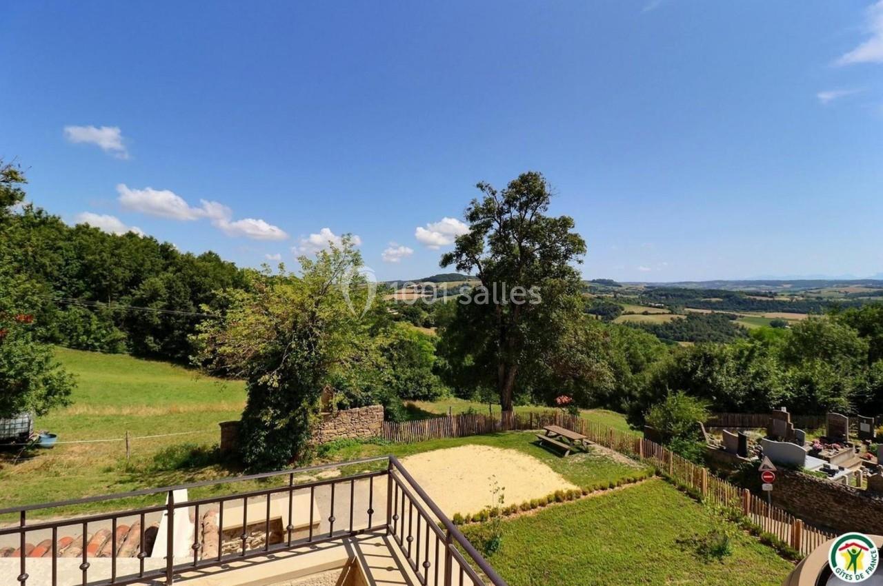 Vue depuis une terrasse sur un paysage rural avec prairies, arbres, collines et un espace aménagé en contrebas.