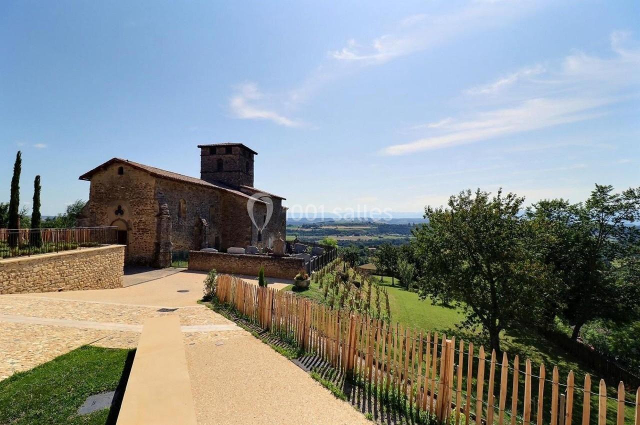 Église en pierre entourée de verdure avec une vue dégagée sur la campagne sous un ciel bleu.