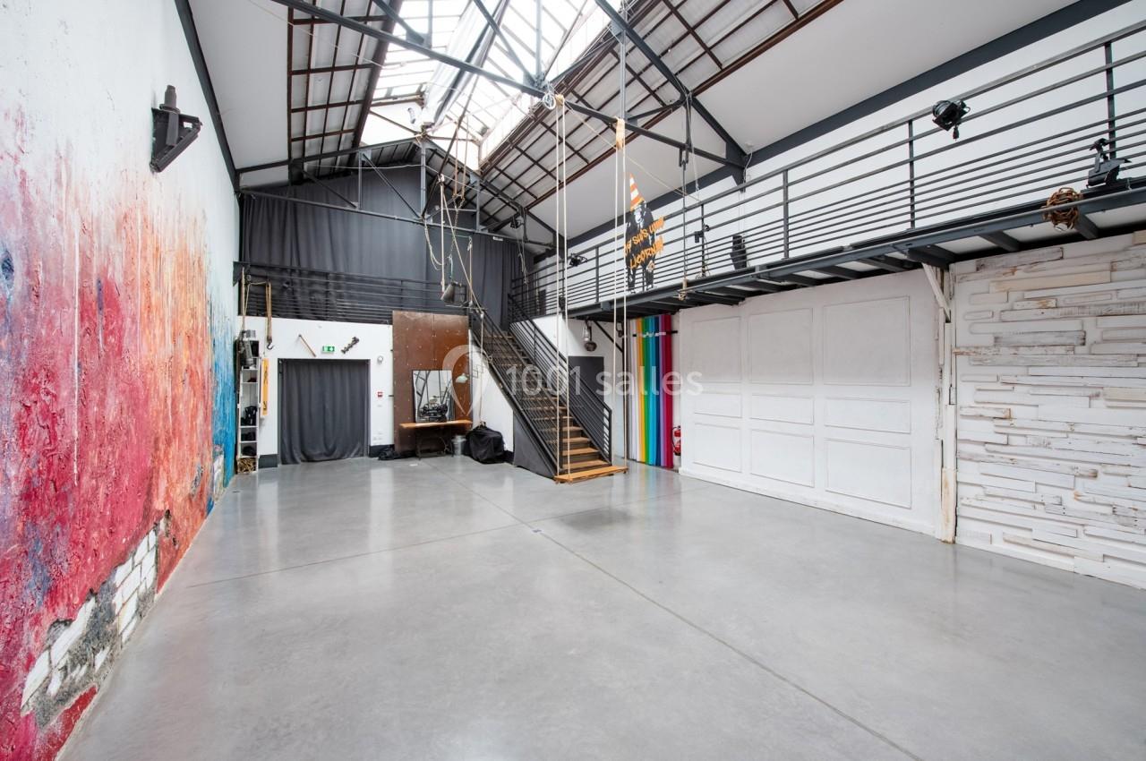 Intérieur d'un loft industriel avec murs colorés, sol en béton, mezzanine et grande verrière laissant entrer la lumière.