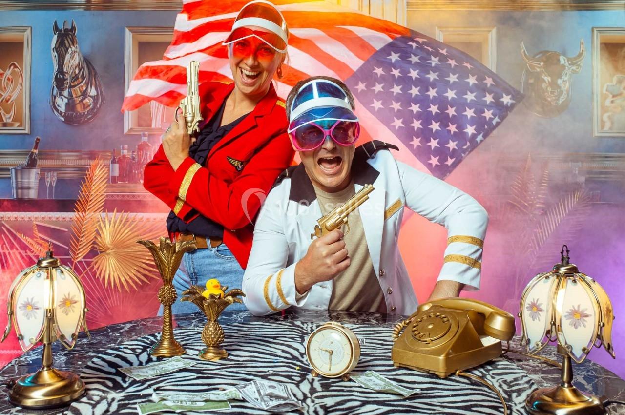 Deux personnes en tenues colorées posent avec des pistolets factices devant un décor kitsch et un drapeau américain.