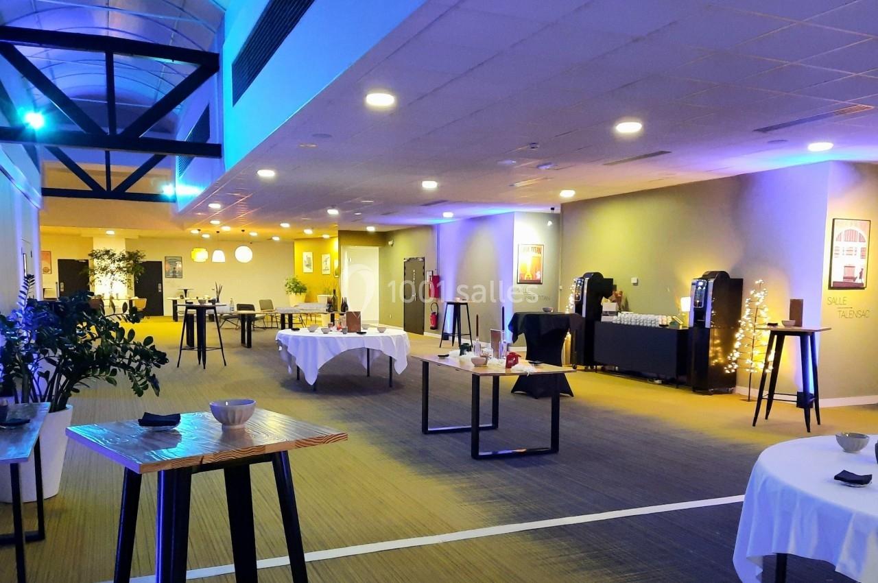 Salle lumineuse aménagée pour un événement, avec tables hautes, nappes blanches et éclairage tamisé.