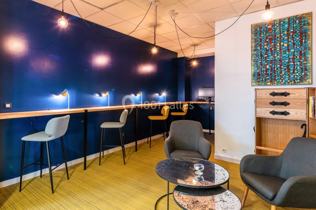 Espace de travail moderne avec murs bleus, chaises hautes, fauteuils et éclairage suspendu.