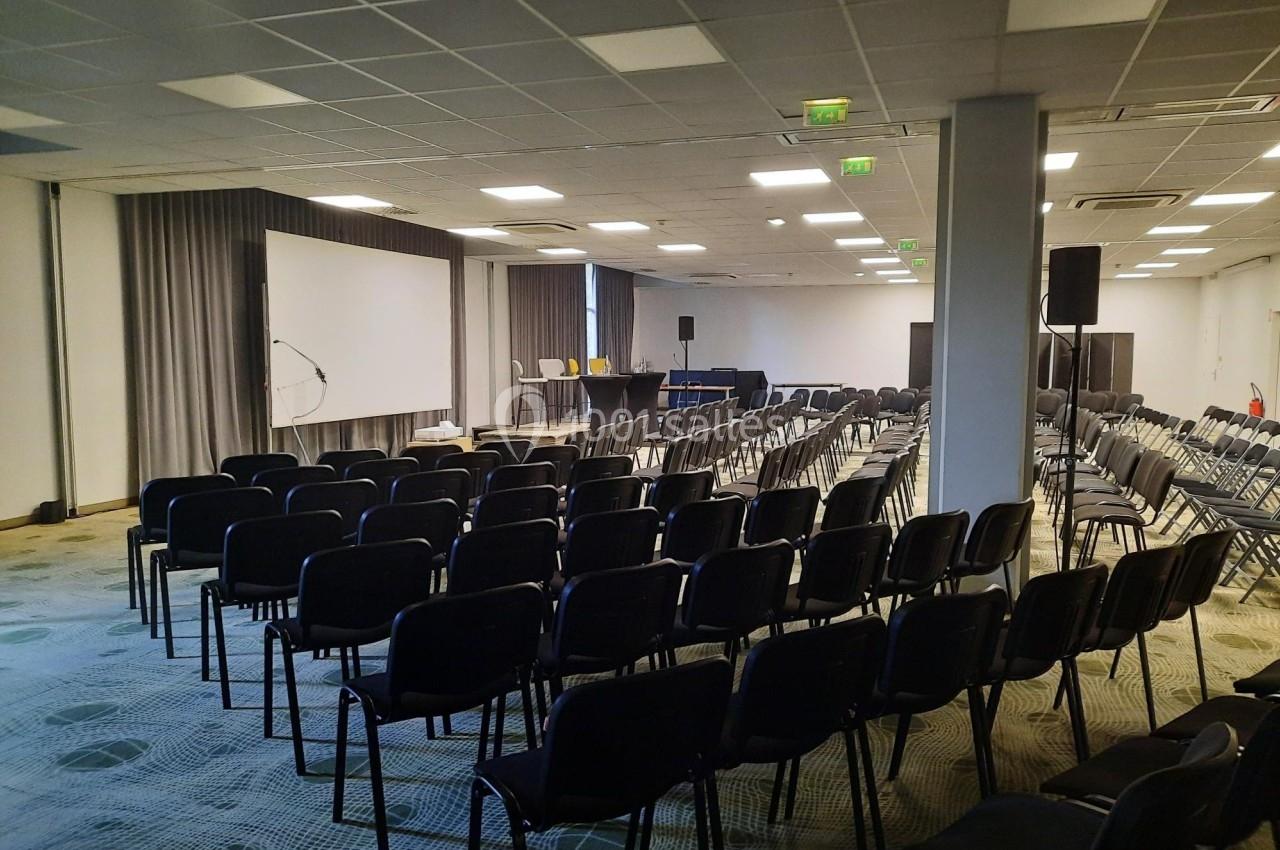 Salle de conférence vide avec des rangées de chaises noires, un écran de projection et un pupitre à l'avant.
