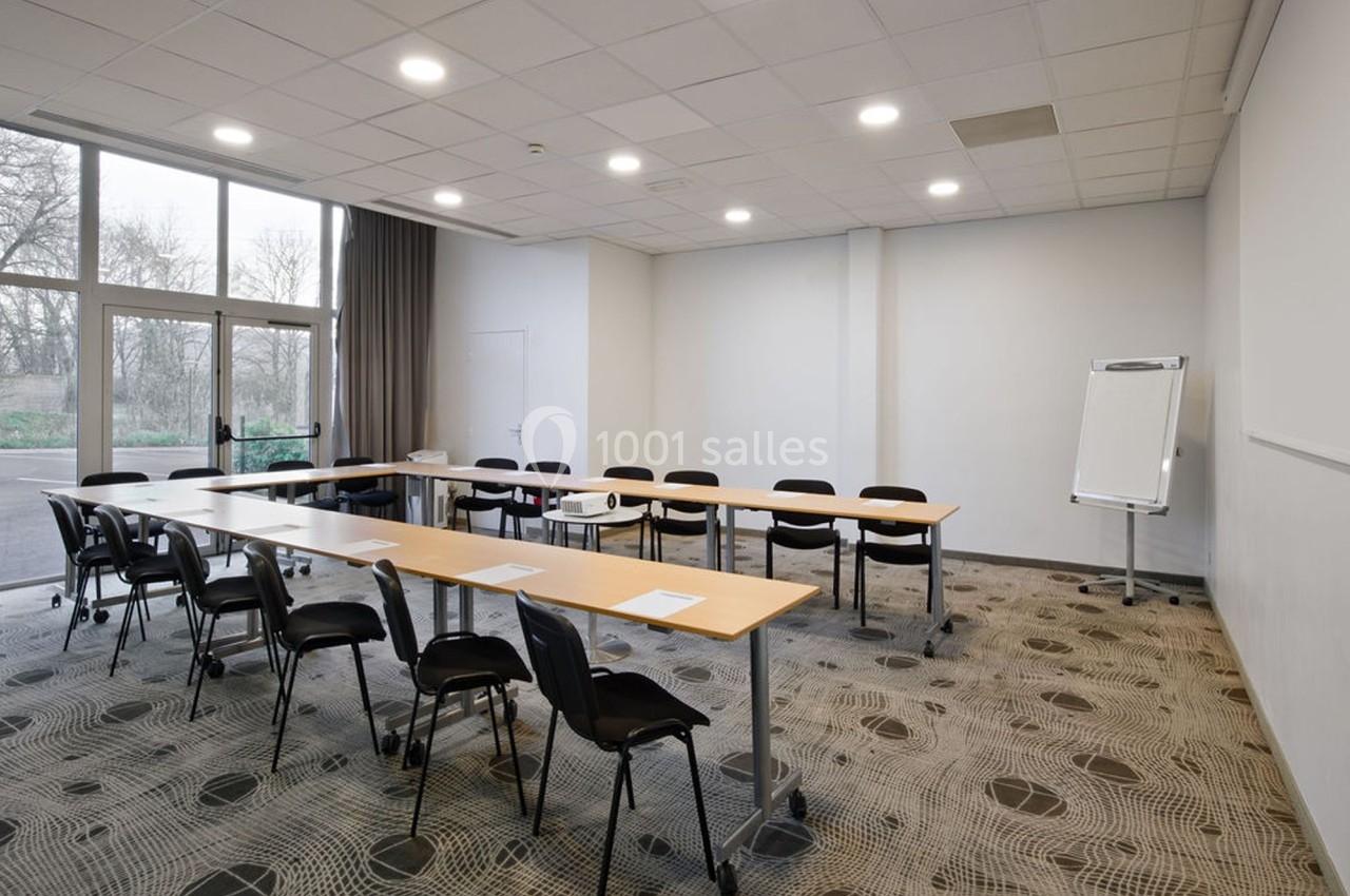 Salle de réunion lumineuse avec tables disposées en U, chaises noires, tableau de conférence et grande baie vitrée.