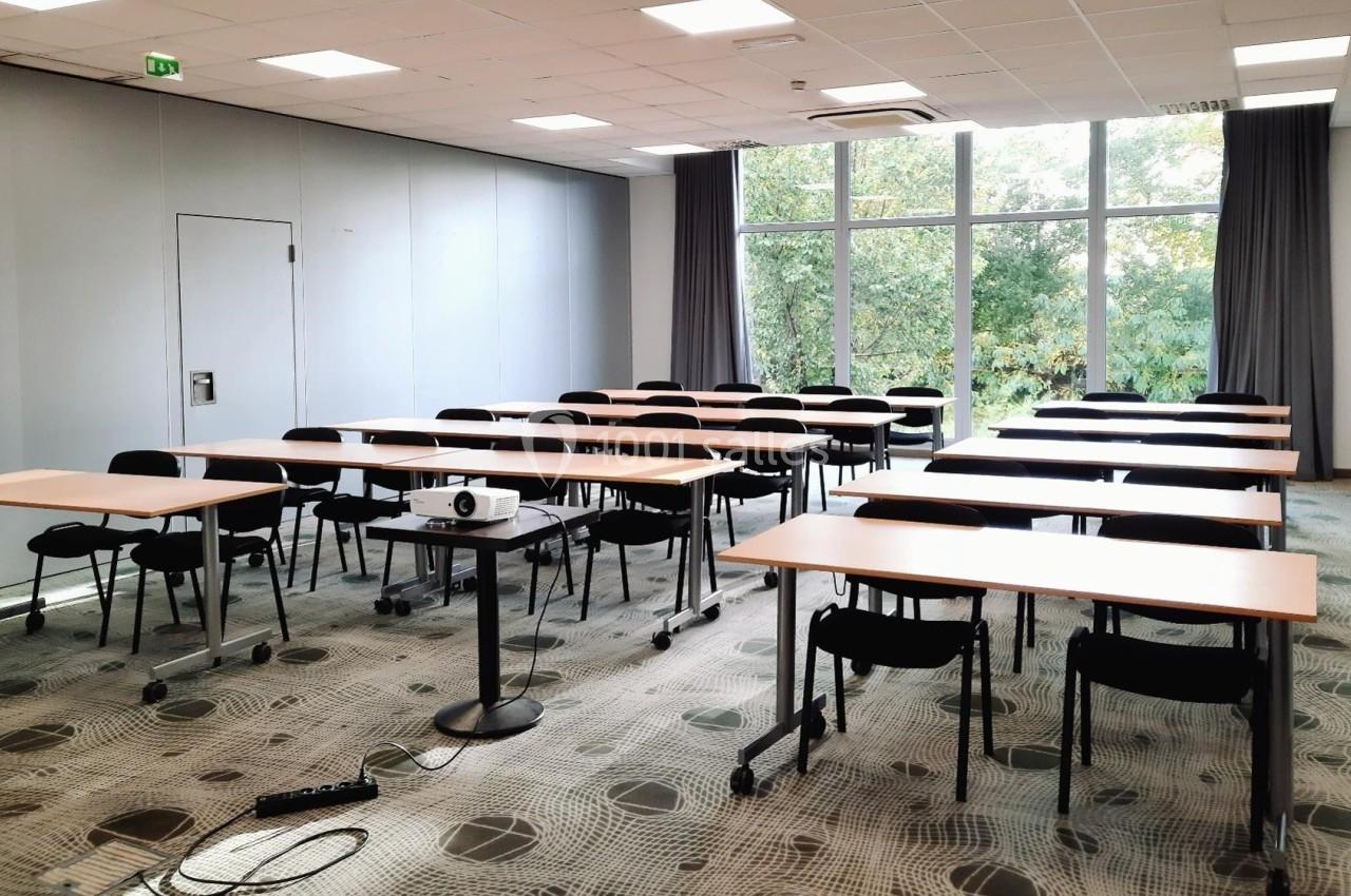 Salle de réunion lumineuse avec des tables et chaises alignées, équipée d'un projecteur et donnant sur un espace arboré.