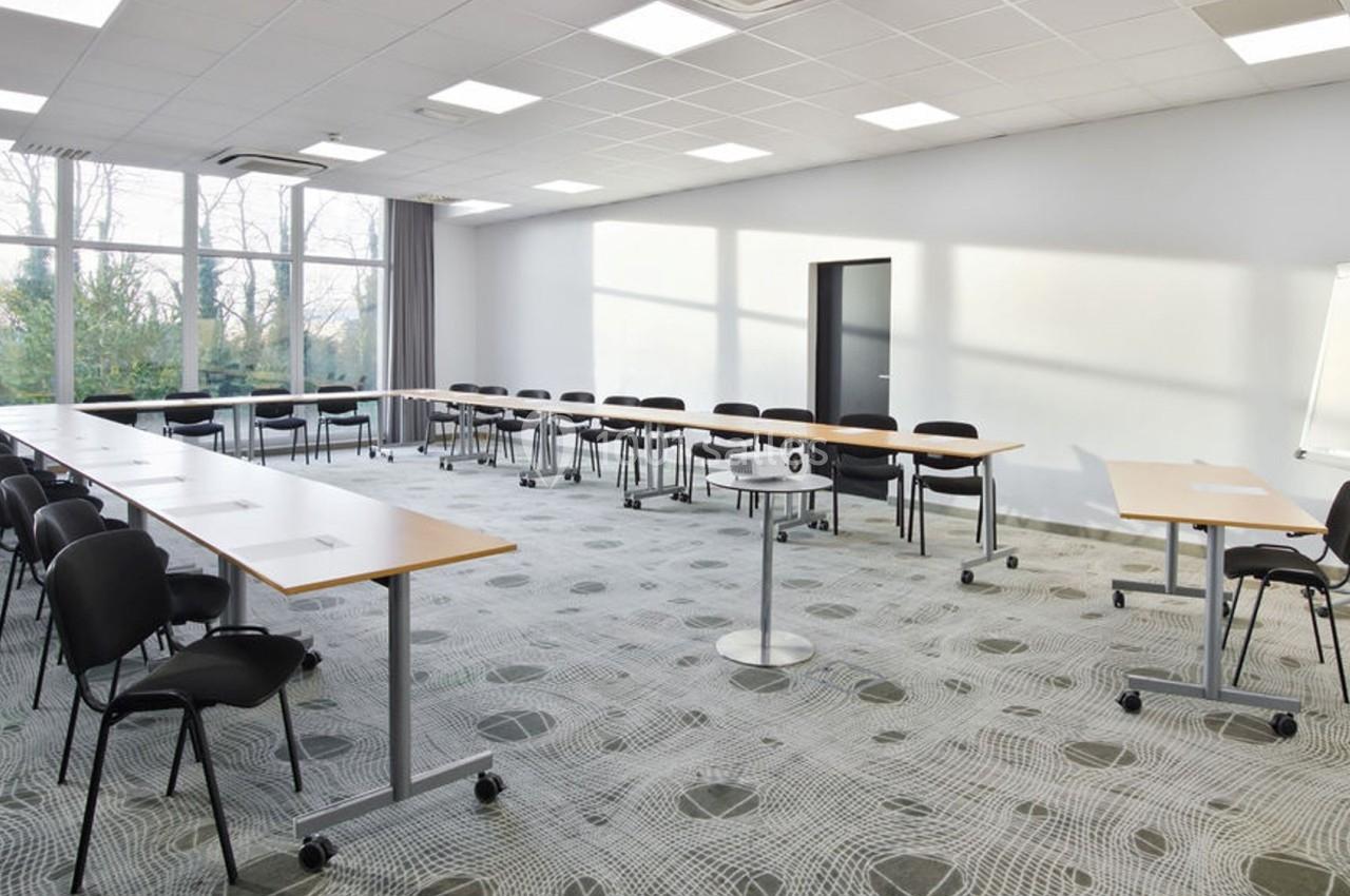 Salle de réunion lumineuse avec tables disposées en U, chaises noires, tableau blanc et projecteur au centre.