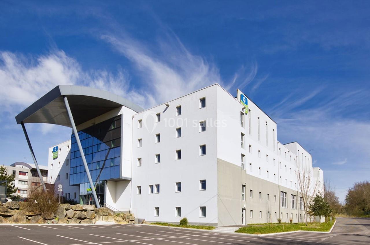 Bâtiment moderne avec façade blanche, grandes fenêtres et parking à l'avant, sous un ciel bleu dégagé.