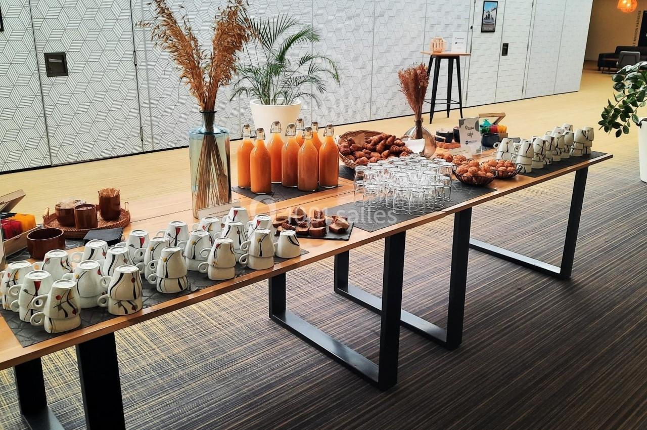 Table de buffet avec jus d'orange, viennoiseries, tasses, et décorations végétales dans un espace lumineux.