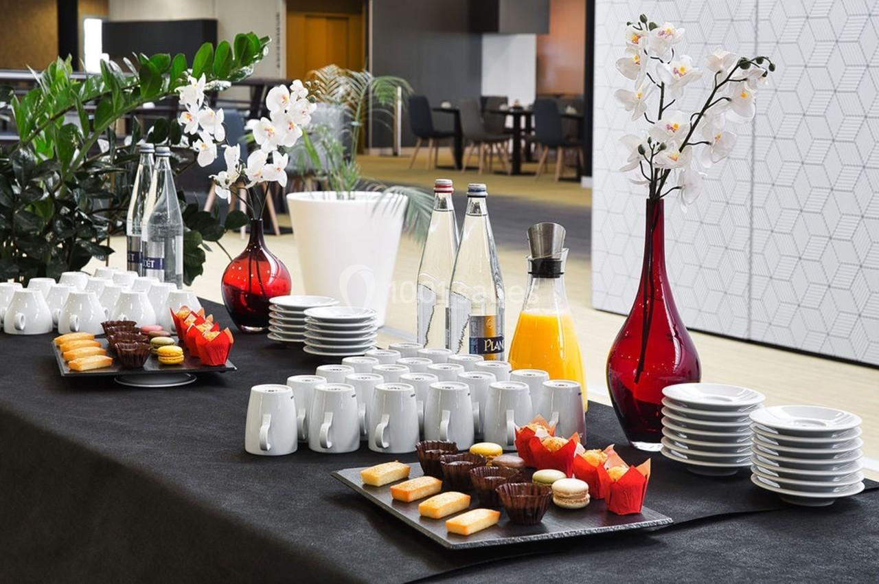 Table avec service de boissons, viennoiseries et vaisselle, décorée de plantes et de vases rouges dans un espace intérieur.