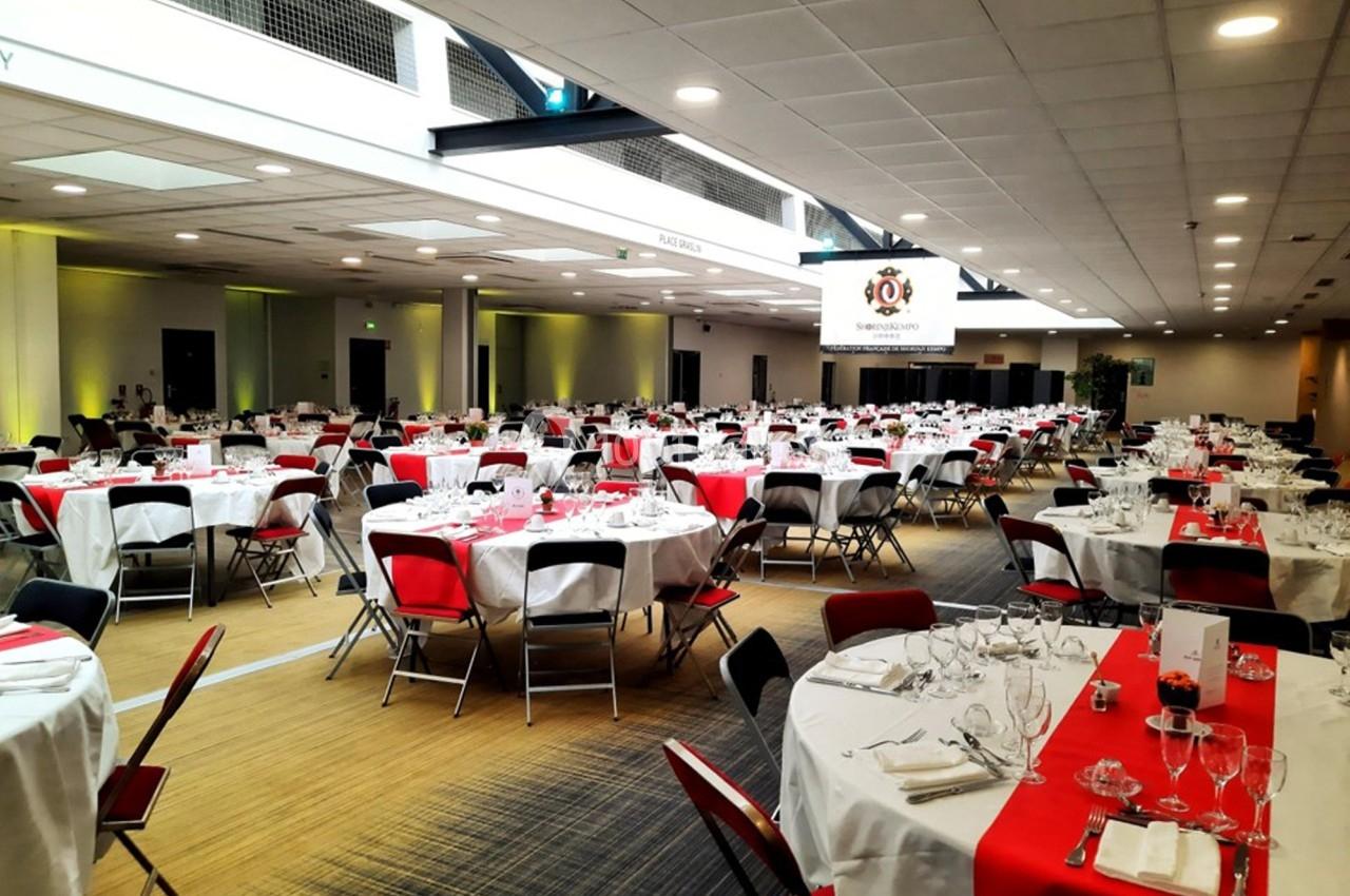 Salle de réception avec tables rondes dressées, nappes blanches et chemins de table rouges, éclairage intérieur lumineux.