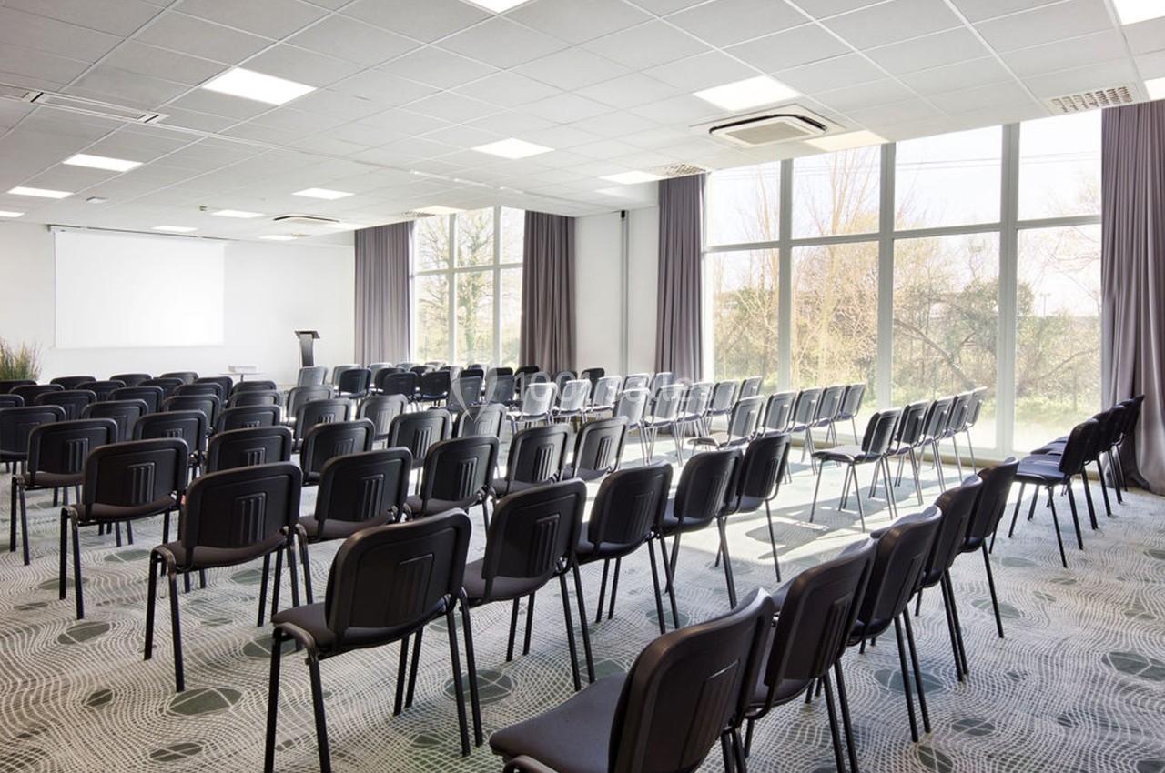 Salle de conférence lumineuse avec des rangées de chaises noires alignées face à un écran blanc et un pupitre.