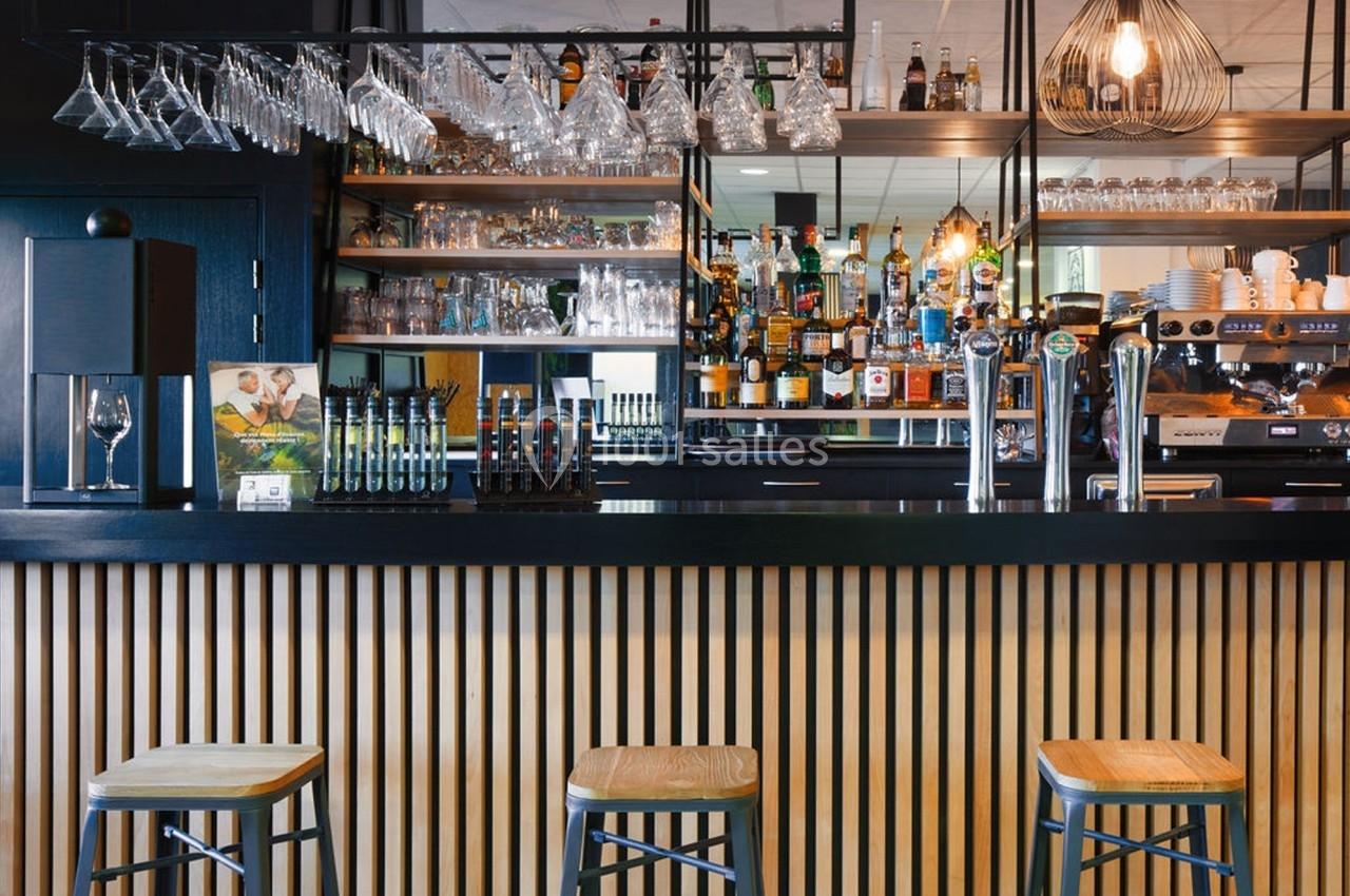 Bar moderne avec comptoir en bois, tabourets, verres suspendus et étagères remplies de bouteilles et tasses.