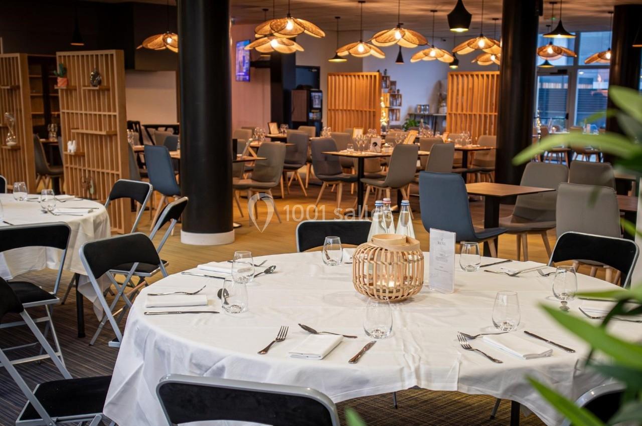 Salle de restaurant avec tables rondes dressées, chaises noires et grises, éclairage chaleureux et décoration moderne.